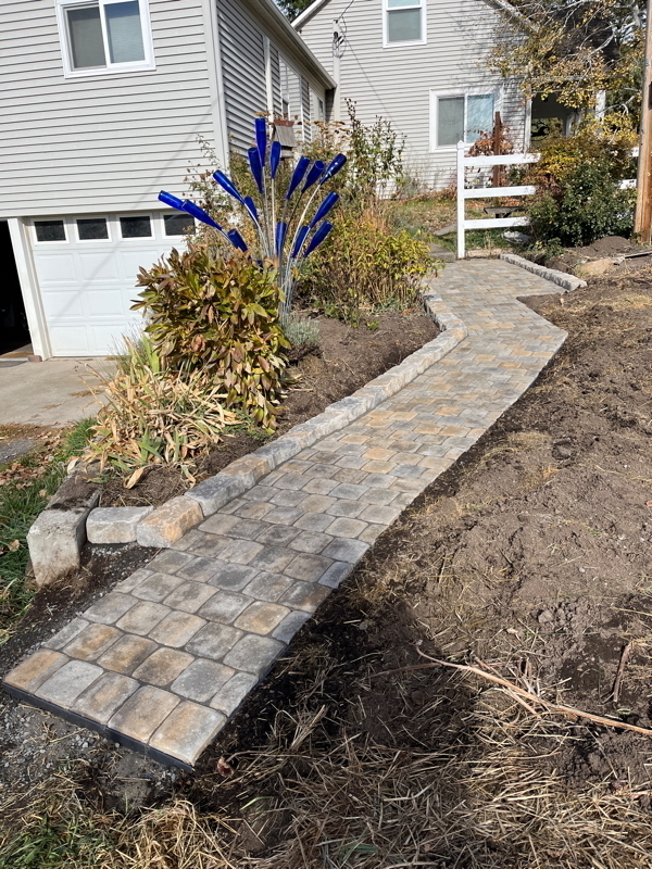 Auto-generated description: A newly constructed stone path winds through a garden area beside a house with a visible driveway and garage.